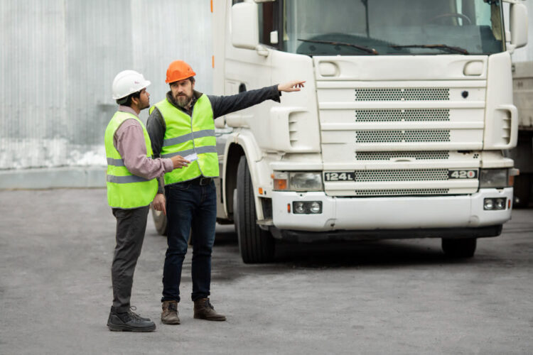 Two construction workers in safety vests and hard hats discussing logistics beside a large truck at an industrial site